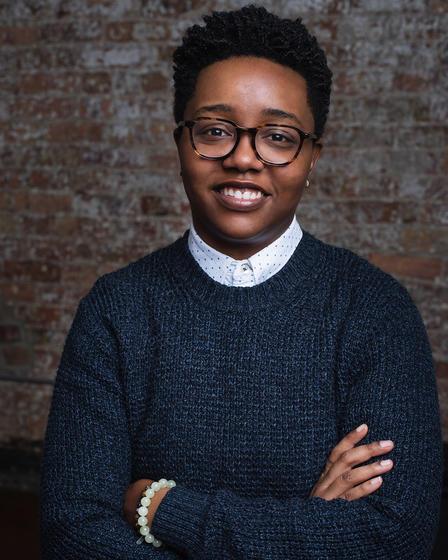 A headshot of Shá Norman, the Director of Diversity for the College of Visual and Performing Arts. They are a Black person with short natural hair, wearing tortoiseshell framed glasses, and smiling at the camera with their arms folded low across their stomach. They are wearing a dark grey sweater with a collared shirt underneath.
